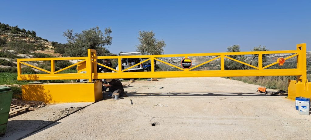 שער מתכת צהוב גדול מותקן על דרך. השער עשוי ממסגרת מתכת עם דפוס צולב, ועליו לוגו "פנחס היישריק בע"מ". ברקע ניתן לראות שטח פתוח עם עצים ונוף הררי.