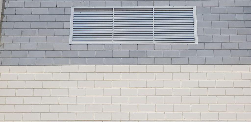 קיר בנוי מאריחים בגווני אפור ולבן, ובחלקו העליון מותקנת רפפת מתכת עם תריסים אופקיים.