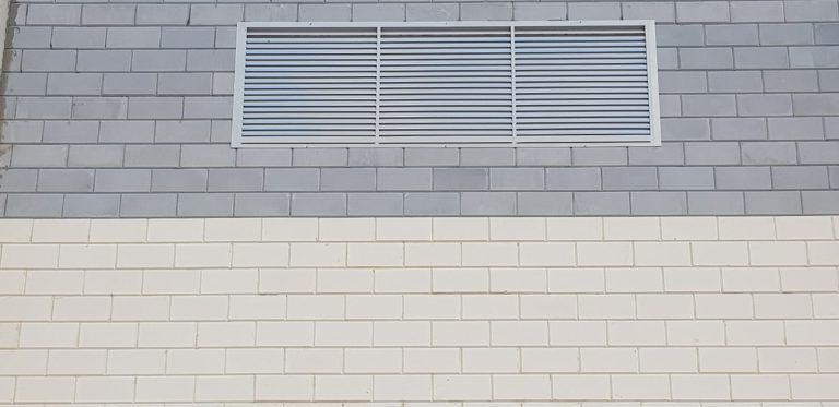 קיר בנוי מאריחים בגווני אפור ולבן, ובחלקו העליון מותקנת רפפת מתכת עם תריסים אופקיים.