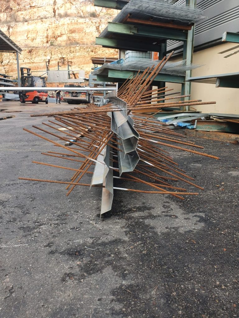 קורות מתכת מגולוונות במבנה זוויתי עם מוטות ברזל חלוד היוצאים מכל צד. הקורות מונחות על הקרקע באזור עבודה חיצוני, וברקע ניתן לראות מדפים עם ציוד מתכתי נוסף.