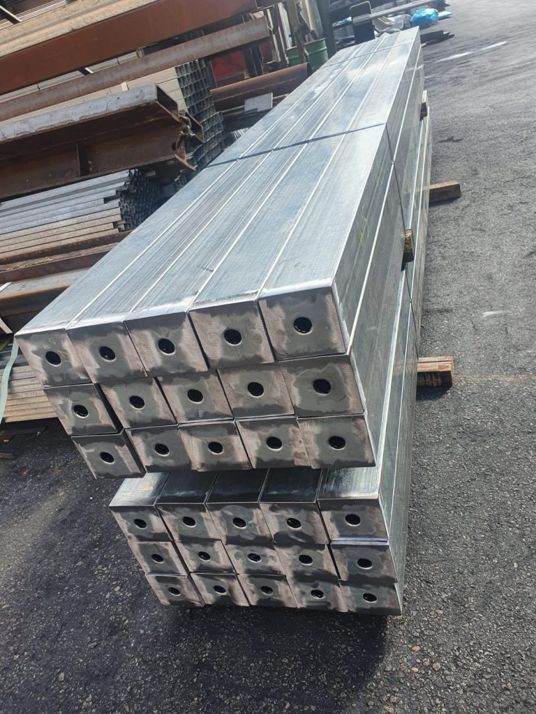 ערימה של פרופילי ברזל מרובעים מגולוונים עם חורים בקצוותיהם, מסודרת על גבי קרשים באתר עבודה חיצוני.