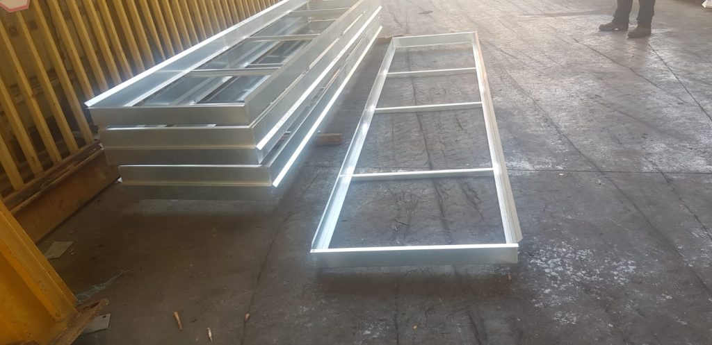 מסגרות ברזל מגולוון מונחות בערימה על רצפת בטון, כאשר אחת מהן מונחת בצד. ברקע קיר עץ מחורץ.