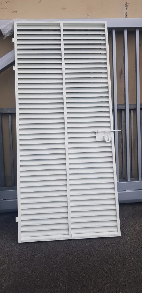 דלת רפפות מתכת לבנה עם תריסים אופקיים, מונחת בזווית על הקרקע. מאחוריה נראה אלמנט מתכת נוסף בצבע אפור.